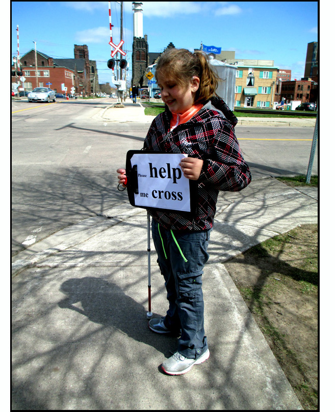 The youngest student stands at the corner.  She is smiling and holding up a clip board with the 8.5 x 11-inch sign that says 'please HELP me to CROSS.'