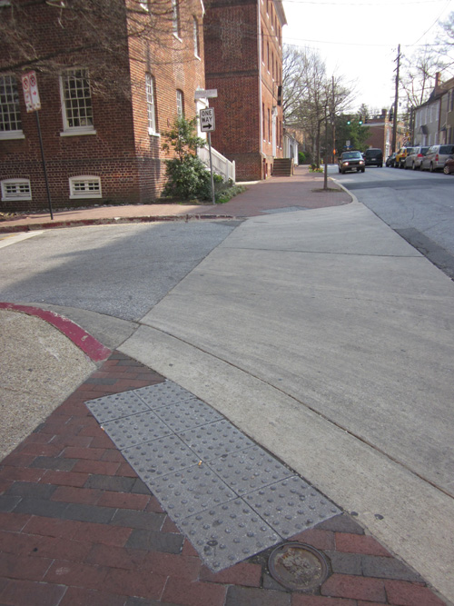 Photo shows a curbramp where the sidewalk is made of red bricks but the sidewalk nearby as well as the street are made of concrete.  Two rows of 4 one-foot-square concrete detectable warning tiles are placed along the edge of the curbramp.