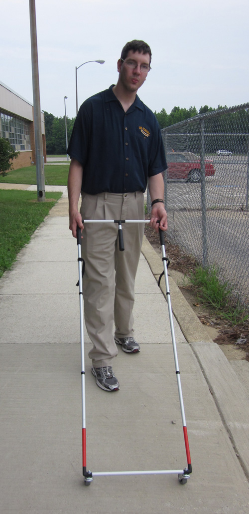 Photo shows a young man walking along a sidewalk pushing another Ambutech AMD.  It is rectangular with a swivel wheel at the bottom corners, and all white except for about 4 inches of red at the bottom of the sides.