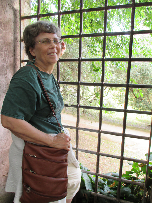 Photo shows Dona smiling at the camera and facing a gate that looks into trees and bushes.