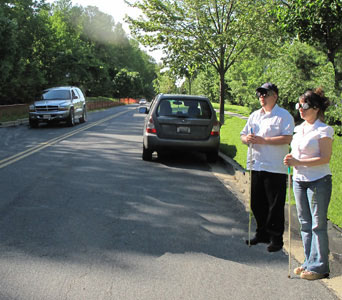 Photo shows Paul and Jomania standing at a curb with a vehicle parked to their right.  Further to their right a car is approaching them.