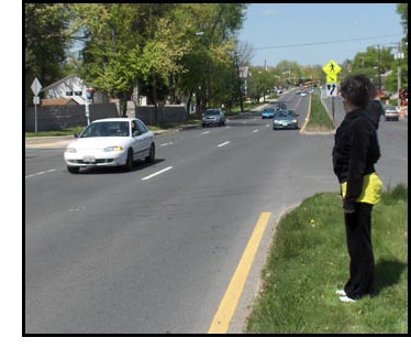 Two pictures show a woman standing on a wide grassy median facing 3 lanes with no stop sign or traffic signal.  The first picture shows no approaching traffic visible from her right for about two blocks, the second shows one car in the second lane in front of her and other traffic approaching in the other lanes.