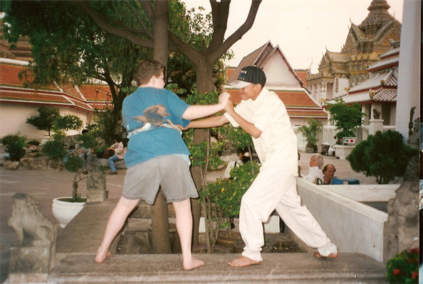 Two teenage boys -- Nanta's son and Stephan, a boy with curly light hair -- stand on a wall and are grabbing each other's hands and pushing eachother, grinning.