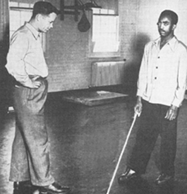 in a large empty gymnasium, Stanley Suterko stands with hands on hips and watches a black soldier hold a white cane, which appears to be long enough to reach his elbow from the floor.