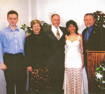 On the left is Stephan, who is tall and wearing a blue button-down shirt, next is Dona wearing a brown blazer and long skirt, then is Paul wearing a suit and tie and holding his arms around Dona and his bride Jomania who wears a white wedding dress with straps, the bottom of the skirt is white translucent lace.  She is standing next to Fred who wears a dark suit and tie.  All are smiling.  Green spring wreathes are on the white wall behind the wedding party and on the podium beside them.