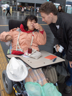 3 photos show Stephan and a young Japanese woman in electric wheelchair with tray.  She has her arms raised about chest-high with her hands clenched and she is leaning far back, saying something while smiling at Stephan