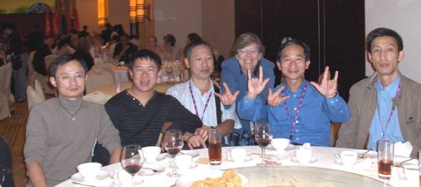 Five men sit at the table while Dona stands behind them -- she and David are making the 'I-love-you' sign