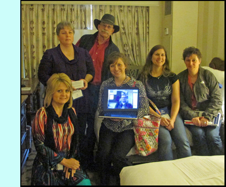 photo shows the members sitting on a bed in a hotel room, Sarah is holding a laptop with Daniel shown on the screen