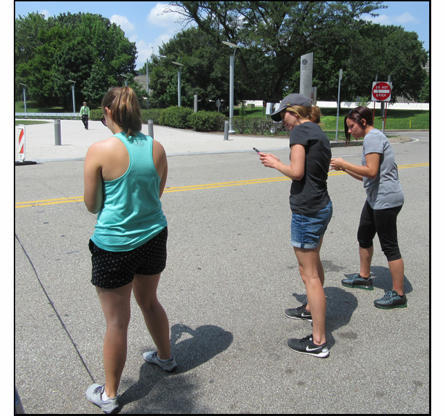 Photo shows 3 women standing at the edge of a two-lane street, holding timers and looking to the left.