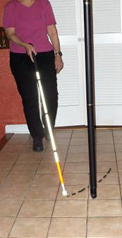series of photos shows a woman walking toward a pole with a cane in her right hand.  the cane tip starts on the woman's left, misses the pole as it moves to the woman's right, and when it reaches left again the entire cane is beyond the pole and the woman bumps into the pole with her left shoulder.