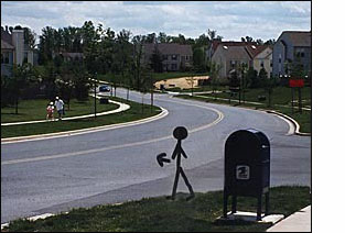 The figure/person on the corner faces a two-lane street. To the right of the figure (east) the street is straight and goes slightly downhill for about 50 feet and then curves to the left and is visible going straight uphill for another half block.