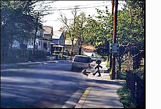 The figure/person on the corner faces a street with two moving lanes plus enough room for cars to park on both sides. To the right of the figure (north) the street is straight for about a block, and has a minivan and other vehicles parked along the street, close to the corner.