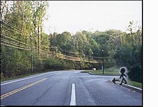 The figure/person on the corner faces a 3-lane residential highway with thick woods on both sides. About a half block to the right of the figure (west) the street makes a sharp turn to the right and disappears behind the trees.