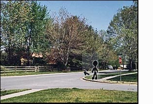 The figure/person on the corner faces a two-lane residential street. To the right of the figure (east) the street is straight only for about half a block and then it turns sharply to the left and is no longer visible. A van appears as it comes around the bend.