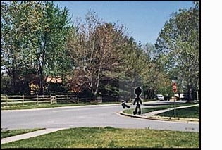 The figure/person on the corner faces a two-lane residential street.  To the right of the figure (east) the street is straight only for about half a block and then it turns sharply to the left and is no longer visible.  A van appears as it comes around the bend.