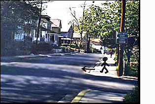 The figure/person on the corner faces a street with two moving lanes plus enough room for cars to park on both sides. To the right of the figure (north) the street is straight for about a block. There are no cars parked in the street.