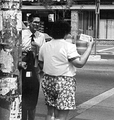 Photo shows the back of a woman with white short-sleeved shirt and flowered shorts standing at the crosswalk, with the card help over her right shoulder.  A man wearing a long-sleeved white shirt and tie and carrying a newspaper and his suit coat is approaching from her left, looking very concerned.