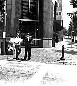 Picture shows the same two men as the last photo. They are both facing the street and the deaf-blind man with the white shirt is starting to walk, holding his white cane in his right hand and reaching for the other man with his left hand. The man in the suit is beside him and holding his left elbow, looking down.