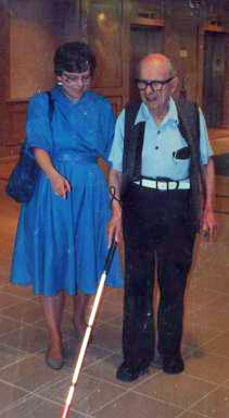 A visually impaired elderly man wearing glasses holds a cane while Dona talks to him and points to the cane.