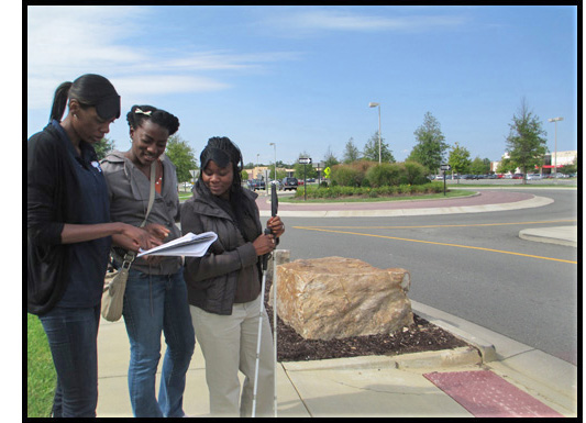 3 participants stand at a crosswalk and check their notes -- one of them is holding a cane and wearing a blindfold which has been lifted so she can see the notes. They are standing at a crosswalk, and to their left we can see a roundabout.