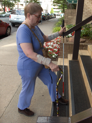 Photo shows woman at bottom of stairs, with the support cane in her right hand and long white cane in her left hand, both canes are held vertically.  Her right leg is still on the ground and her left leg and both canes are on the first step.