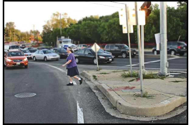 Photo shows a person crossing a lane of right-turning traffic, starting from an island and going to the curb.  A car in the lane is waiting for her to cross -- on the other side of the island are 3 lanes of traffic waiting for the traffic signal.