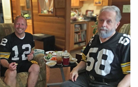 Paul and Fred are wearing Steeler shirts and sitting in a living room with snacks.  Paul is looking at us and grinning very broadly, and Fred is watching the game.