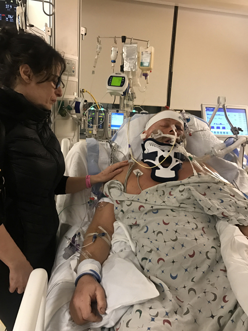 Picture shows Paul unconscious in hospital bed, head bandanged with breathing tube and lots of wires and tubes from his head, one leg wrapped.
