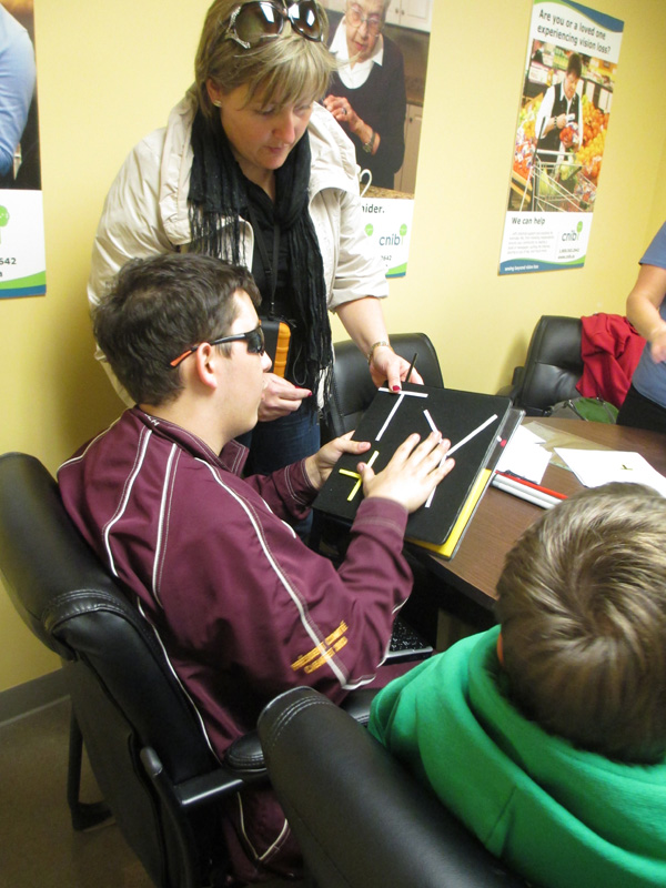 These 4 photos show students sitting around a large conference table with instructors standing and talking, or walking around the table to show a tactile map of intersection shapes to several of the students.