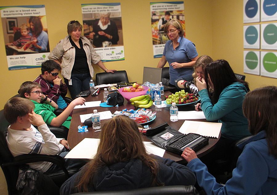 These 4 photos show students sitting around a large conference table with instructors standing and talking, or walking around the table to show a tactile map of intersection shapes to several of the students.