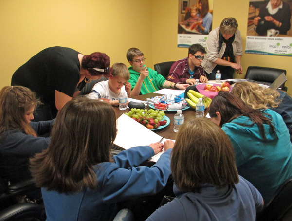 These 4 photos show students sitting around a large conference table with instructors standing and talking, or walking around the table to show a tactile map of intersection shapes to several of the students.