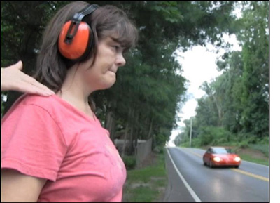 picture shows a woman with headphones facing a two-lane street where a car is approaching close to her left, and someone is tapping her on her shoulder.