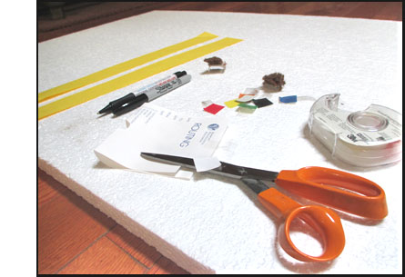 Photo shows a white foam board about 3'x3' with a pen and a marker, tape, two strips of yellow paper about 2 inches wide and 2 feet long, a half-dozen one-inch-square pieces of paper of various colors, and a scizzors cutting another one-inch-square piece of white paper.