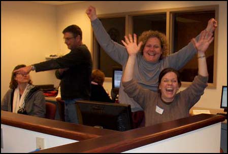 Photo shows a row of computers, with one woman sitting in front of a computer and another woman standing behind her, both of them laughing and triumphantly throwing their hands up.  Other women sit at other computers and a man points to something on one of their screens.