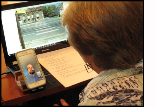 Picture shows Dona sitting at her computer, with a picture of a crosswalk on the screen and a checklist on the keyboard.  She is looking at Facetime on her smartphone, where she can see her student Jeremy.