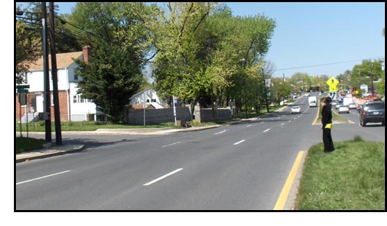 A woman stands on a wide grassy median facing 3 lanes of traffic coming from her right -- behind her is the other half of the street.  There is traffic approaching from about a block away on her right.  There is no traffic signal at her intersection, there is a stop sign for the street beside her but not for the street she is facing.