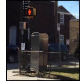 Photo shows a street corner with a pedestrian signal and pushbutton suspended on a telephone pole.  Sitting on the grass nearby is a large metal box about 5 feet high, 3 feet wide and 2 feet deep.
