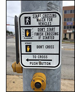 Photo shows two pedestrian pushbuttons on a wide metal pole, one button is facing us and the other is facing to our left.  Each has a sign above it.  The top of the sign shows a silhouette of a walking pedestrian and says 'START CROSSING - watch for vehicles.' The middle shows a flashing orange hand and says 'DON'T START.  Finish crossing if started.'  Near the bottom is an orange hand and says, 'DON'T CROSS.'  At the bottom is an arrow pointing toward the street to our left and says, 'TO CROSS, PUSH BUTTON.'   