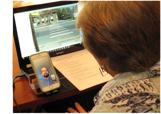 Picture shows Dona sitting at her computer, with a picture of a crosswalk on the screen and a checklist on the keyboard.  She is looking at Facetime on her smartphone, where she can see her student Jeremy.