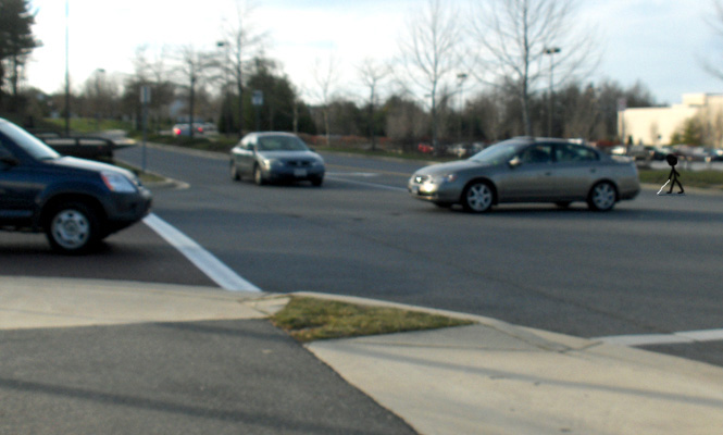 Photo shows a car that is half-way across a 3-lane street, and a stick figure with a cane is starting to cross to the right of where the car had been waiting at a stop sign.
To the right of the car is another car approaching along the main street, about 10 feet away from the first car.