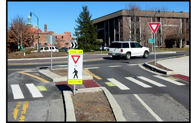 Photo shows a roundabout, starting with two-lane street with a crosswalk that cuts through a median strip.  A sign in the median strip says 'yield to pedestrians.' About 10 feet past the crosswalk, there is a 'yield' sign where street merges into a circular roadway that goes around a large island. 