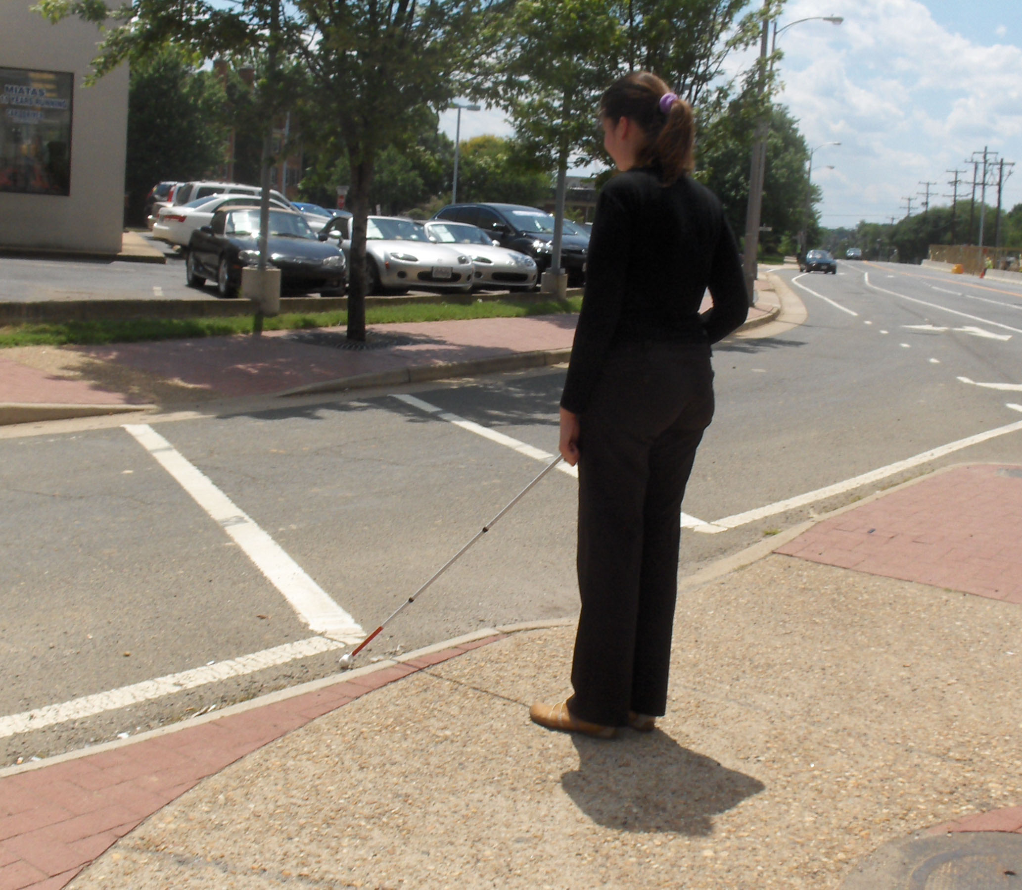 Two photos show Mimi standing on the SW island with her cane near the crosswalk of the right-turning lane.  To her right is a street with one or two cars.  The second photo shows a minivan leaving the street and turning slightly to its right to enter the lane in front of Mimi and approach the crosswalk.