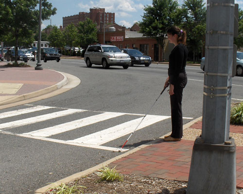 Two photos show Mimi standing on the SE island with her cane in the crosswalk of the right-turning lane.  To her right is a very busy street.  The second photo shows a car turning right from the busy street almost 90 degrees into the lane in front of Mimi and approaching the crosswalk.