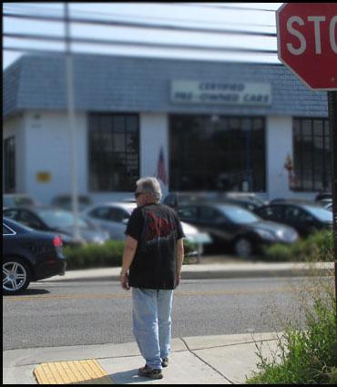 Photo shows a man standing at a corner facing a street.  there is a stopsign for the street beside him but not for the street he is facing.