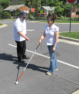 Photo shows student with opaque goggles walking with a cane across the lanes in the parking lot.  The instructor is pointing to her foot which is at the end of the first lane.
